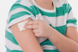 Imagem mostra uma criança colocando a mão no ombro com um curativo depois de tomar a vacina.