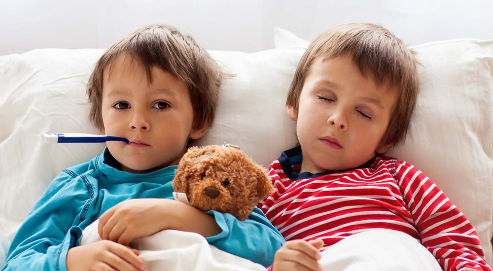 Duas crianças sentadas na cama. Uma está com um termômetro na boca e o ursinho no braço. Ambas com sintomas de febre amarela.