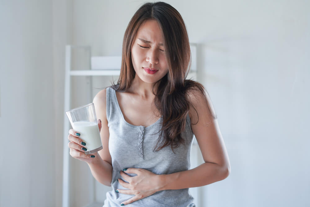 Uma mulher com alergia alimentar segurando um copo de leite com uma mão, e com a outra pressionando a barriga demonstrando estar sentindo dor
