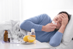 Um homem doente está deitado assoando o nariz. em frente tem uma mesa com alguns remédios