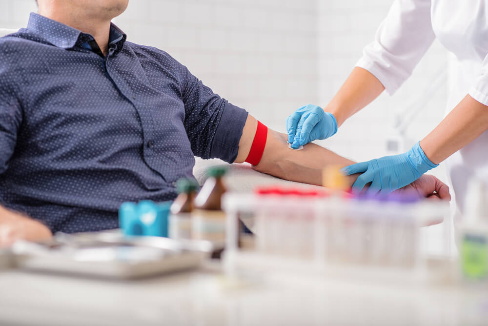 Um homem está sentado e com o braço esticado, enquanto a médica está fazendo a higienização na região onde será feita a coleta de sangue