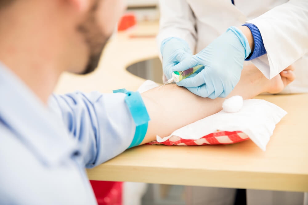 Um homem com o braço esticado e apoiado em cima de uma mesa, enquanto um médico está fazendo a coleta de sangue