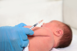 um bebê deitado de lado e a mão de um profissional de saúde se preparando para aplicar injeção