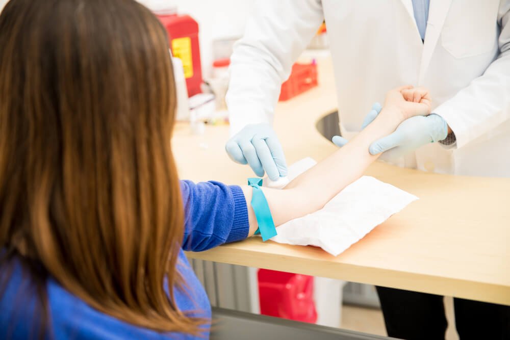 Mulher com o braço estendido em cima de uma mesa com um elástico amarrado no braço. Ao fundo, mostra um profissional da saúde preparando o exame de sangue. A foto serve para ilustrar o post sobre o que é exame de tsh.