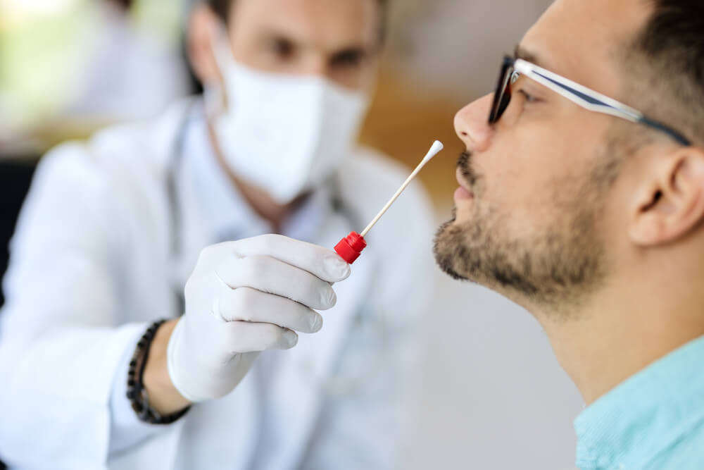 Ao fundo um enfermeiro colocando o swab perto do nariz de um homem sentado de lado durante o exame de PCR para Covid-19.