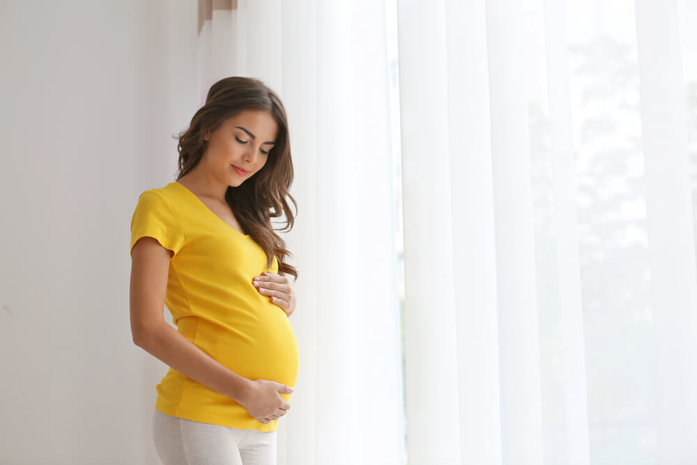 Mulher grávida em pé perto da janela olhando para baixo fazendo carinho na barriga. A imagem ilustra o post sobre qual nome do exame de sangue que detecta gravidez. 