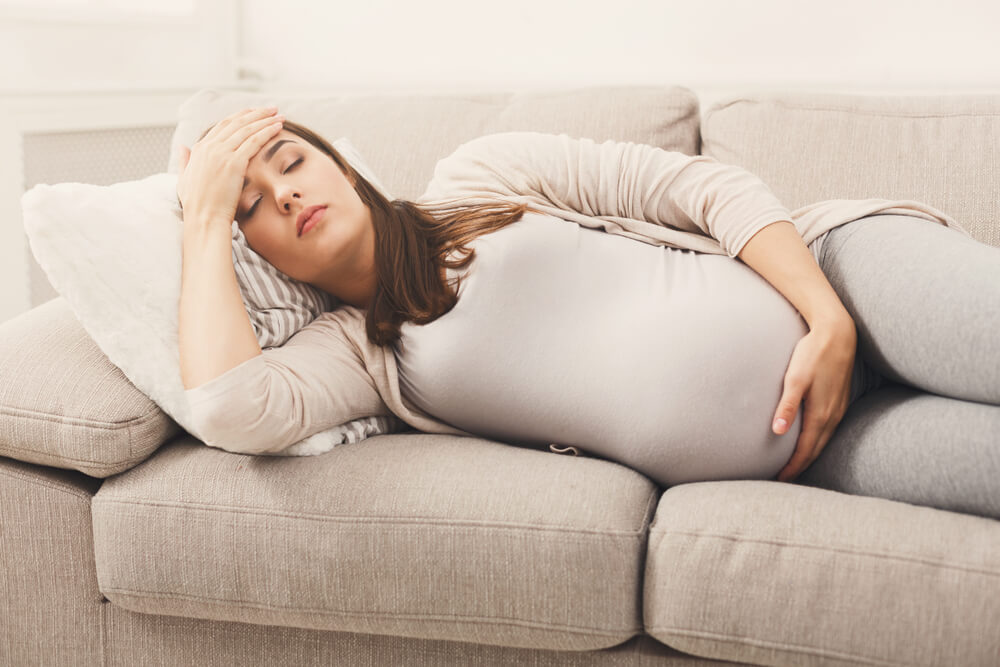 uma mulher gestante deitada de lado em um sofá, com uma mão apoiando na barriga e a outra mão apoiada na cabeça sentindo algum sintoma de gravidez