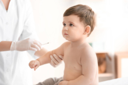Bebê sentado de lado com uma enfermeira segurando o braço com uma injeção na mão para completar o calendário de vacina da criança.