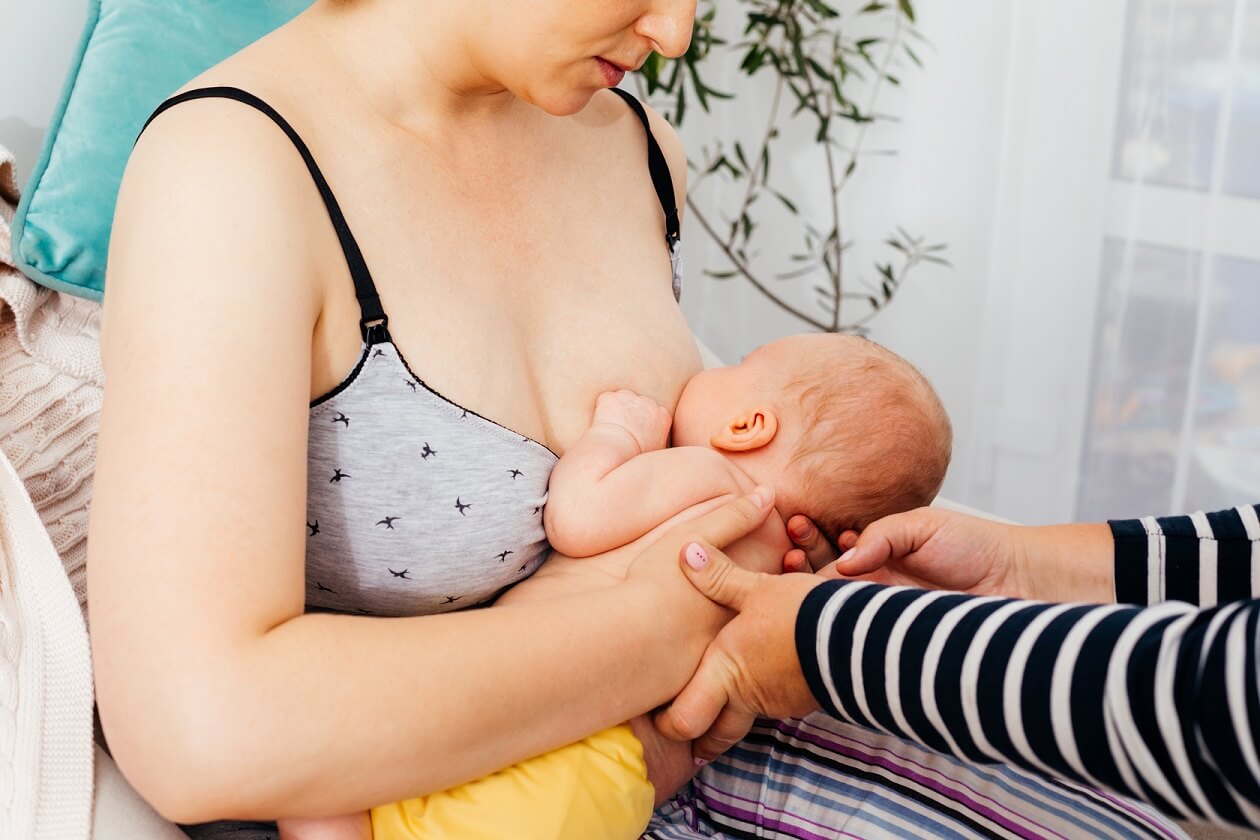 Mulher com o bebê no colo amamentando. As mãos da consultora de amamentação ajudam a segurar a cabeça da criança enquanto o bebê toma o leite materno.