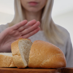 uma imagem mostrando uma mulher parada de frente para uma mesa com pão fazendo sinal de recusa com a mão, demonstrando ter doença celíaca