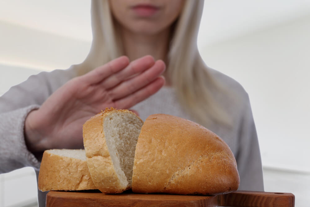 uma imagem mostrando uma mulher parada de frente para uma mesa com pão fazendo sinal de recusa com a mão, demonstrando ter doença celíaca