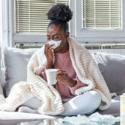 uma mulher sentada no sofá assoando nariz e segurando uma caneca, onde aparenta estar com gripe - perguntas sobre a gripe