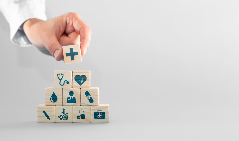 Pilha de cubinhos. Cada um tem um símbolo na frente que representa algo relacionado à saúde. No topo, uma mão coloca o último cubinho. Essa imagem ilustra o post sobre o Dia Mundial da Saúde.