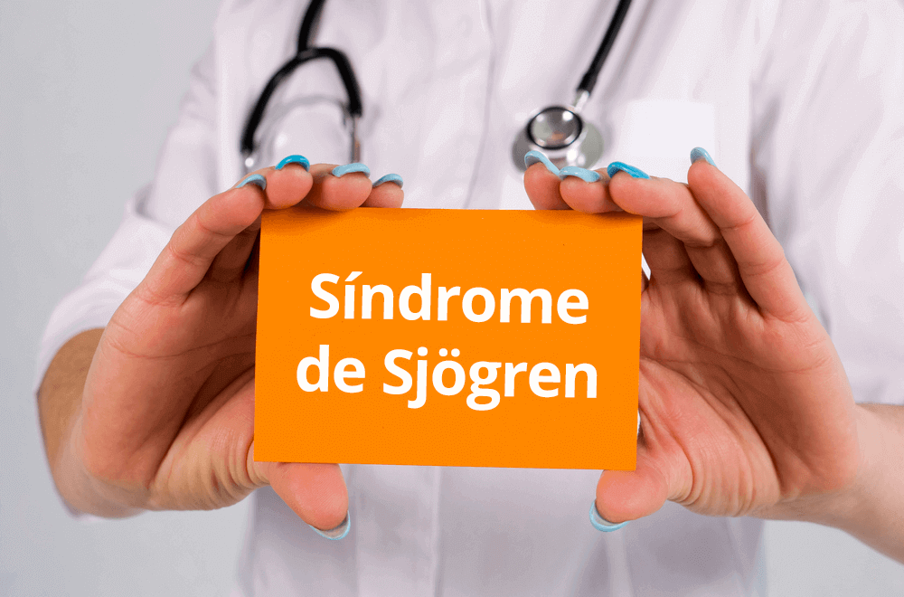 uma imagem mostrando as mãos de um profissional de saúde segurando um cartão coral escrito Síndrome de Sjögren