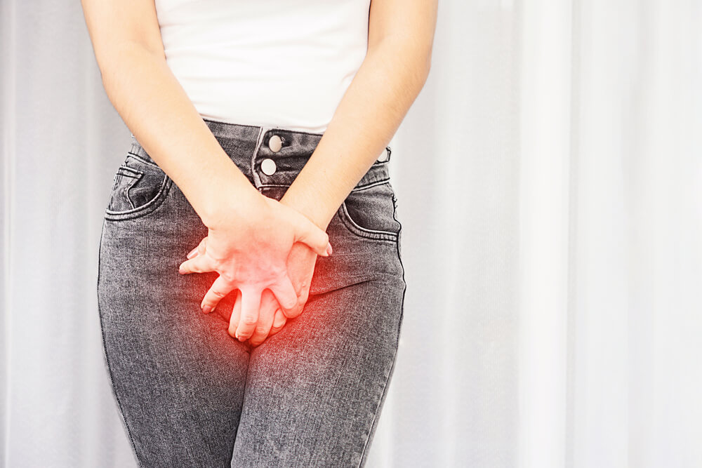 Essa imagem mostra a parte inferior do corpo de uma mulher, que está vestindo calça jeans preta. Ela está cruzando as duas mãos em frente à sua parte íntima, sinalizando algum sintoma dessa infecção. O que é candidíase?