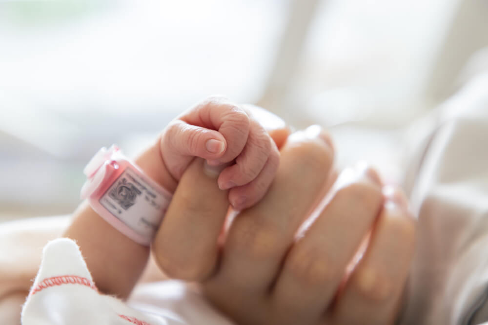 Imagem mostra a mão de um bebê ainda com a identificação do hospital segurando a mão da mãe. Essa imagem representa o post sobre vacina para bebê prematuro. 