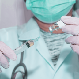 Técnica de enfermagem usando máscara facial segura uma ampola de vacina pneumocócica 13 e passa o líquido para a injeção.