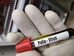 Uma mão com luva branca segurando um tubo de ensaio de tampa vermelha e uma etiqueta escrita pólio vírus