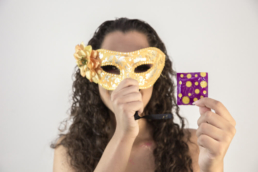 Imagem mostra uma mulher com uma mascara de carnaval no rosto enquanto segura uma camisinha como exemplo de dicas de saude no carnaval