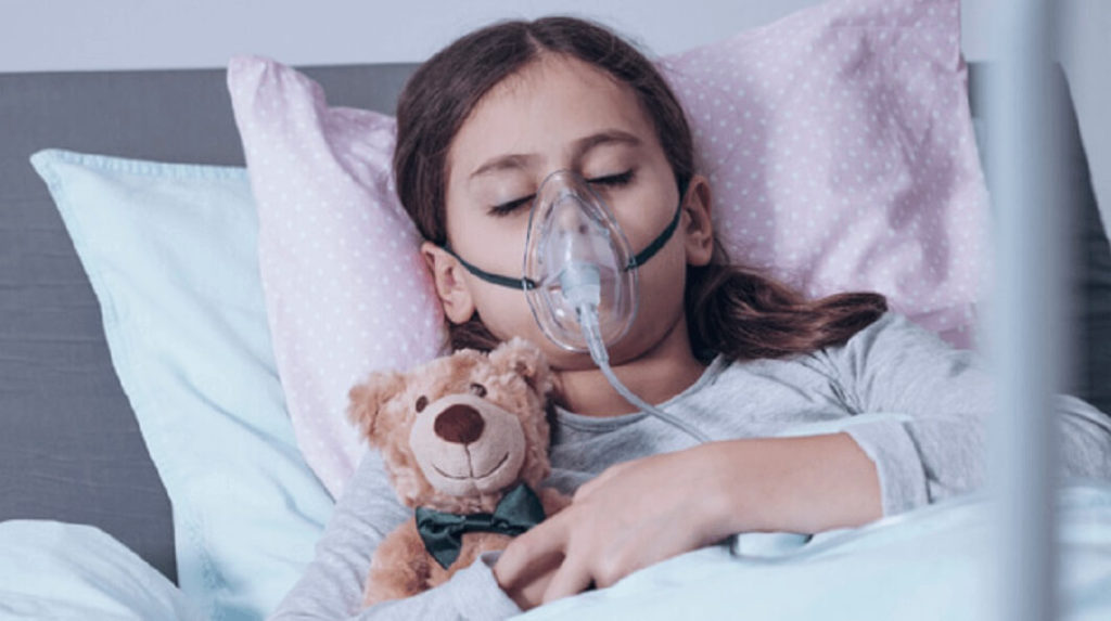 Menina com fibrose cística deitada ao lado de um ursinho de pelúcia e com uma máscara de oxigênio na boca