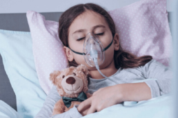 Menina com fibrose cística deitada ao lado de um ursinho de pelúcia e com uma máscara de oxigênio na boca